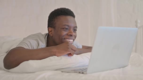 ベッドで胃の上に横たわっている間、若いアフリカ人男性はノートパソコンでビデオチャットを行う - 映像、動画