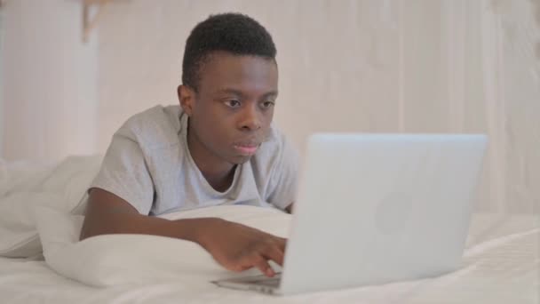 ベッドで胃の上に横たわっている間、ラップトップで働く若いアフリカ人男性 - 映像、動画