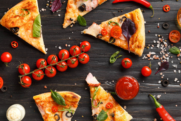 Auf dunklem Holzgrund, einem Zweig frischer Kirschtomaten, Soßen und Gewürzen für die Pizza von oben, liegen Pizzascheiben mit Zitronen, Oliven, Käse und Kräutern. Italienische Lebensmittel. - Foto, Bild