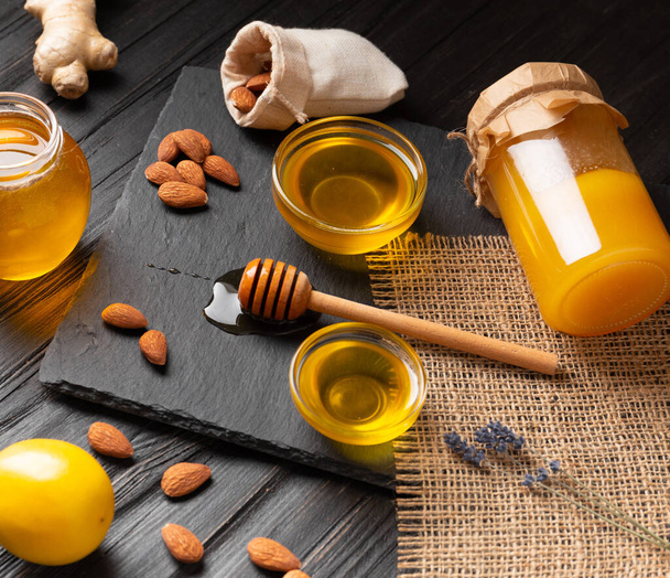 Μέλι σε βάζα, χυλό, λεμόνι, τζίντζερ σε μαύρο πέτρινο πιάτο σε ξύλινο φόντο. Η έννοια των βιολογικών προϊόντων μελισσών. - Φωτογραφία, εικόνα
