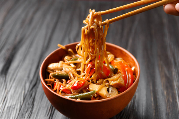 Plate with buckwheat noodles with vegetables, mushrooms, chicken meat and Chinese sticks on a dark background. Japanese soba in clay bowl on a wooden background. - Φωτογραφία, εικόνα