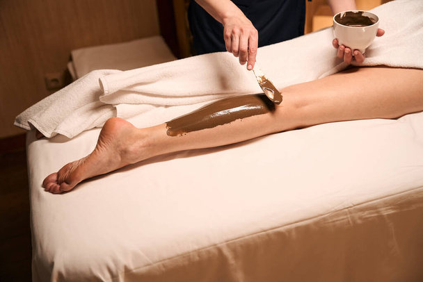 Photo recadrée du dermatologue appliquant de la pâte d'algues sur la jambe du client du spa avec spatule - Photo, image