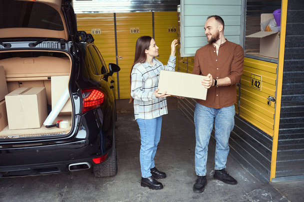 Angenehmer Mann und Frau mit einer Schachtel voller Sachen in der Nähe des Kofferraums eines Autos in einem Abstellraum - Foto, Bild