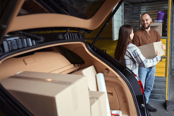 Радісний чоловік і жінка стоять з коробкою в руках біля багажника автомобіля, повного речей
 - Фото, зображення