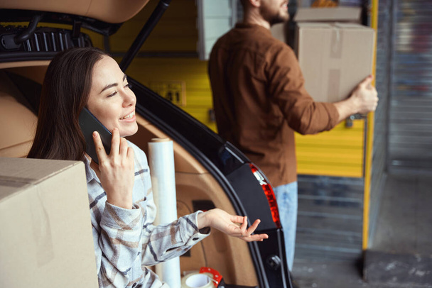 Mujer alegre tener conversación en el teléfono móvil mientras está sentado en el maletero del coche al lado de un hombre trabajador - Foto, imagen
