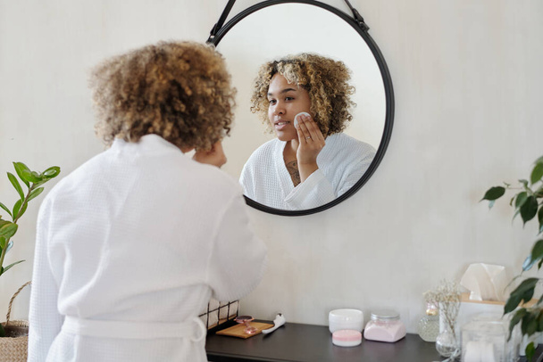 Junge hübsche Frau im weißen Bademantel trägt erfrischendes Toner auf Gesicht mit Wattepad auf, während sie zu Hause vor dem Spiegel steht - Foto, Bild