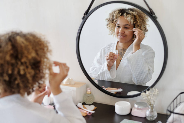 Happy African American dziewczyna stosowania kropli serum nawilżające lub olej na obszarze oczu, stojąc przed lustrem w łazience - Zdjęcie, obraz