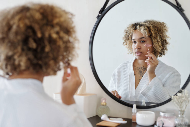 Mladá žena dělá masáž obličeje a nanášení séra na kůži s křemenem váleček, zatímco stojí před zrcadlem v koupelně - Fotografie, Obrázek