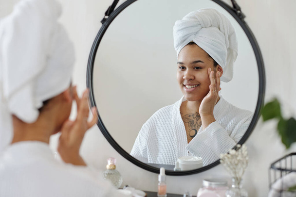 Szczęśliwa młoda kobieta z ręcznikiem na głowie stosując krem nawilżający na twarz patrząc w lustrze w łazience po porannym prysznicu - Zdjęcie, obraz