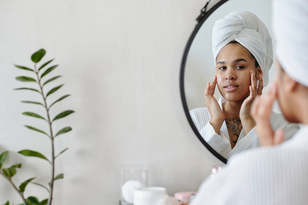 Junge Frau mit Handtuch auf dem Kopf berührt ihr Gesicht nach Schönheitsoperation, während sie im Badezimmer vor dem Spiegel steht - Foto, Bild