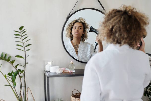 Focus sur la réflexion dans le miroir de la jeune femme noire en peignoir blanc séchant propres cheveux bouclés avec sèche-cheveux électriques dans la salle de bain - Photo, image