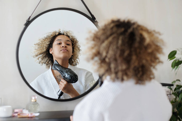 Reflection in mirror of gorgeous African American girl in white bathrobe drying hair with electric hair dryer after washing it with shampoo - Foto, imagen