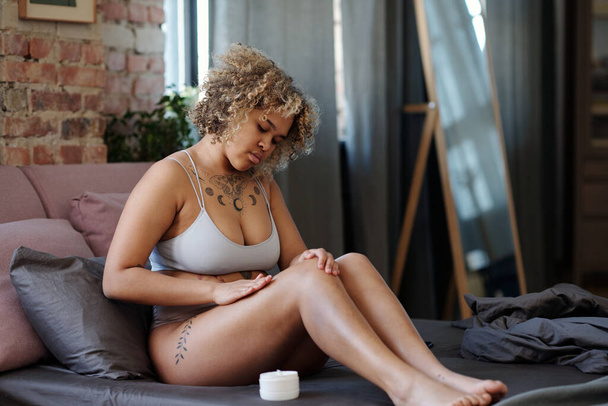 Mujer hermosa joven sentada en la cama y aplicando crema hidratante en la piel de sus piernas después de afeitarlas por la mañana - Foto, imagen