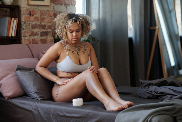 Barefoot African American girl in underwear taking care of leg skin while sitting on bed and applying moisturizing lotion after shaving - Fotó, kép