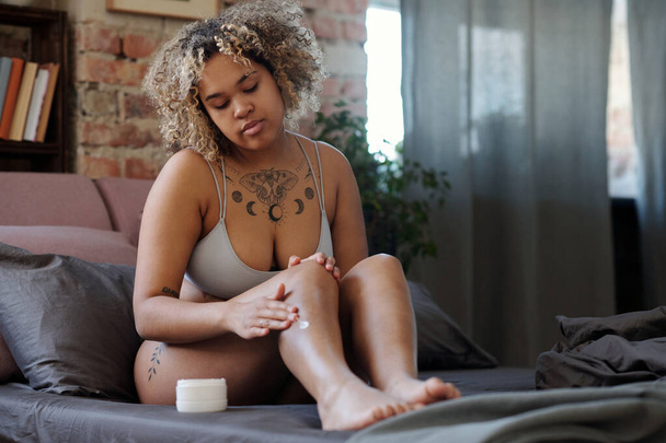 Młoda Afroamerykanka odżywia skórę balsamem do ciała siedząc rano na łóżku po wzięciu prysznica i goleniu nóg - Zdjęcie, obraz