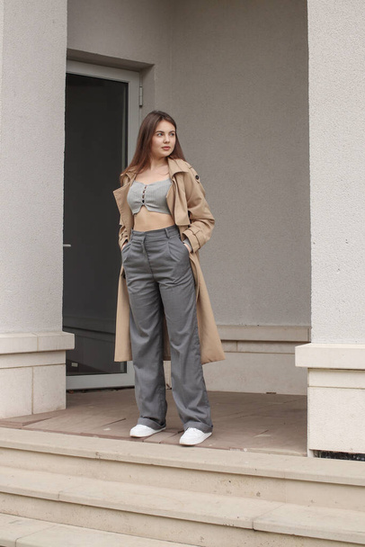 Portrait shooting of a stylish girl. Beige shades. Trends of spring and summer 2023. Classic gray pants and a gray crop top. Trench coat. - Foto, afbeelding
