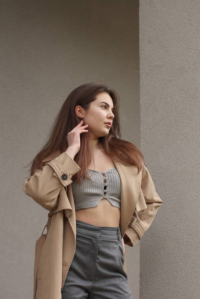 Portrait shooting of a stylish girl. Beige shades. Trends of spring and summer 2023. Classic gray pants and a gray crop top. Trench coat. - Фото, изображение