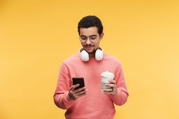Young multi-ethnic man with white headphones around neck using smartphone and having cup of coffee against yellow background - Valokuva, kuva