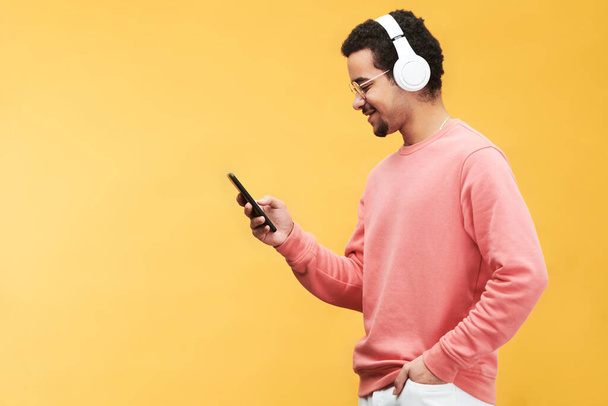Widok z boku szczęśliwy młody wieloetniczny człowiek ze smartfonem słuchanie muzyki w słuchawkach i przewijanie listy odtwarzania w izolacji - Zdjęcie, obraz