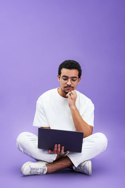 Молодой серьезный человек с ноутбуком на коленях смотрит онлайн видео или работает над новым бизнес-проектом, сидя перед камерой - Фото, изображение
