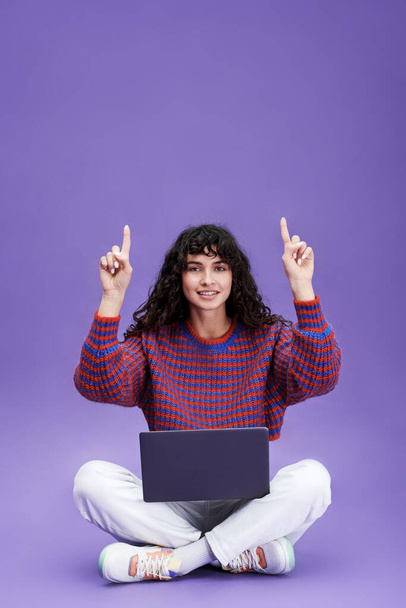 Mujer morena joven feliz con el ordenador portátil en las piernas cruzadas sentado delante de la cámara y apuntando hacia arriba en copyspace para su texto - Foto, Imagen