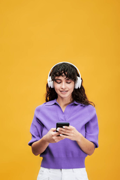 Brunette vrouwelijke student in hoofdtelefoon luisteren naar muziek in de pauze tijdens het scrollen in de afspeellijst of kijken door middel van video 's in smartphone - Foto, afbeelding