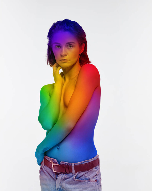 Portrait of young woman standing shirtless with body colored in rainbow colors, symbolizing lgbt acceptance over white background. Concept of lgbt community, support, love, human rights, pride month - Foto, Bild