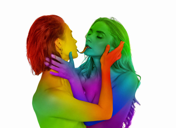 Portrait of loving young women kissing, eating jelly candy against white background. Models painted into rainbow colors. Concept of lgbt community, support, love, human rights, pride month - Foto, imagen