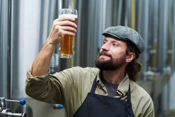 Happy bearded brewery owner looking at glass of beer he made - Foto, Imagem