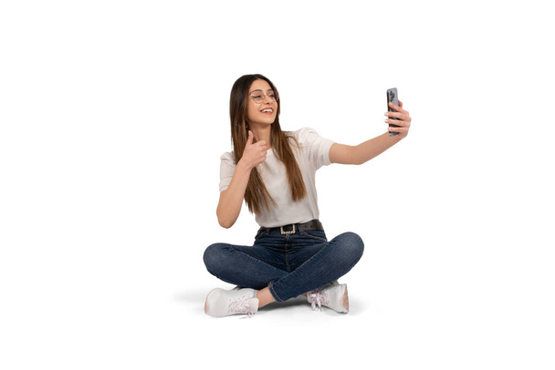 Making a video call, full body portrait of caucasian woman making a video call. Doing ok sign, thumbs up to smartphone camera. Smiling happy woman chatting online with friends. Modern communication. - Фото, изображение
