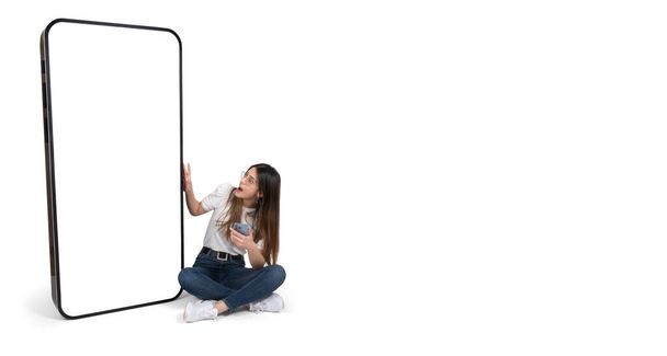 Aufgeregt sitzt eine Frau neben einem großen Smartphone mit leerem Bildschirm-Attrappen. Ganzkörper junge kaukasische Mädchen suchen große Handy und berühren. Mobile Anwendung, Website empfehlen Bannerdesign-Konzept. - Foto, Bild