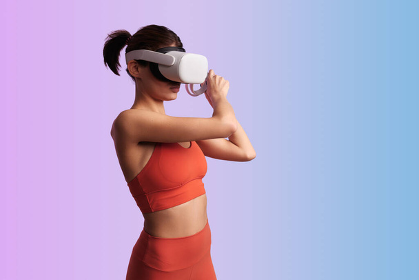 Side view of fit young woman in sportswear and VR headset doing exercise with simulator while standing on colorful gradient background - Φωτογραφία, εικόνα