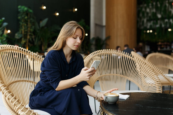 Charming woman chatting on cellphone with cup of aromatic coffee at cafe place. Attractive young lady having conversation on mobile phone near window indoors. Smiling  - Photo, image