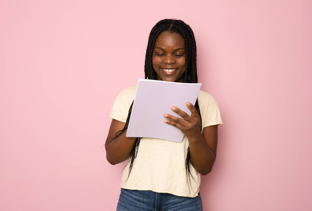 若い笑顔幸せなアフリカ系アメリカ人の少女ティーン学生はピンク色の背景スタジオの肖像画に隔離された本を保持します。高等学校大学構想における教育. - 写真・画像
