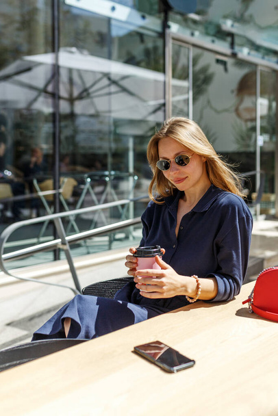 Uma jovem garota hipster bonita com xícara de bebida está sentada em uma mesa de uma cafeteria de verão de rua..  - Foto, Imagem