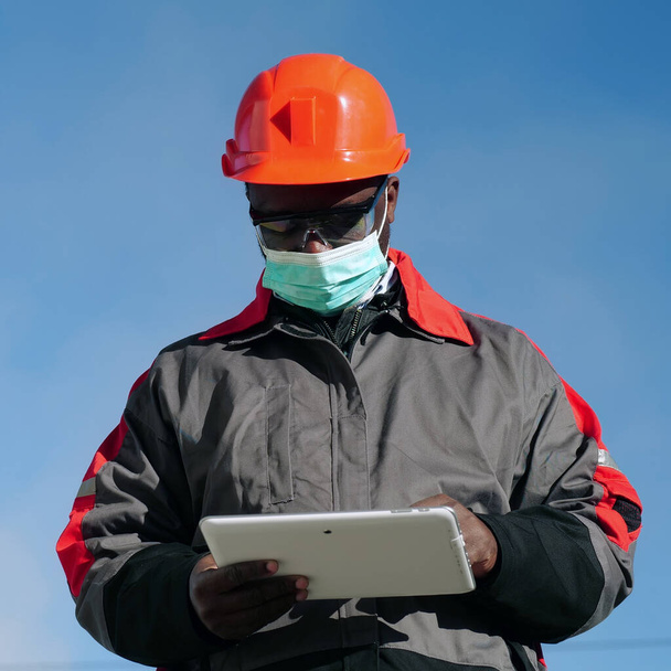 Αφροαμερικάνος εργάτης με μάσκα προσώπου ιατρικής προστασίας στέκεται στο εργοτάξιο και κρατάει σημειώσεις στον υπολογιστή του. Εργάτης κρατά στα χέρια λευκό δισκίο PC - Φωτογραφία, εικόνα