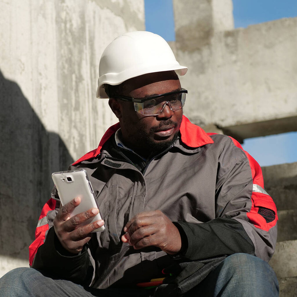 Αφροαμερικανός εργάτης με σκληρό καπέλο και προστατευτικά γυαλιά με smartphone κάθεται στο εργοτάξιο. Εργάτης κατέχει στα χέρια λευκό smartphone και σκέφτεται - Φωτογραφία, εικόνα