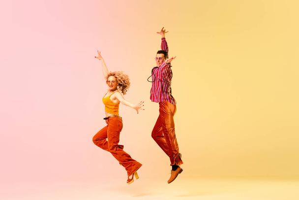 Dynamic image of beautiful young couple, man and woman in stylish vintage clothes dancing against gradient pink yellow background. Concept of retro style, dance, fashion, art, hobby, music, 70s - Fotoğraf, Görsel