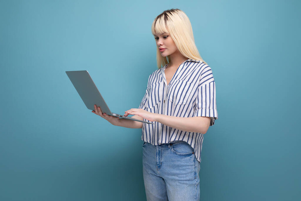 Çizgili tişörtlü ciddi sarışın bir kadın stüdyo arka planında dizüstü bilgisayarın arkasında çalışıyor.. - Fotoğraf, Görsel