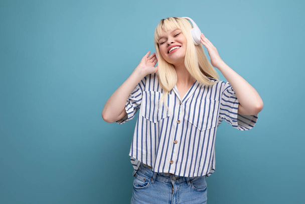 happy blond young woman in striped shirt listening to music with headphones on studio background. - Photo, Image