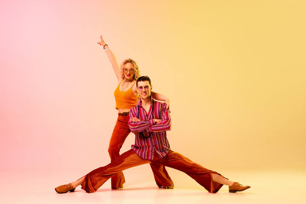 Dynamic image of stylish, talented, emotional couple, man and woman in vintage costumes dancing disco dance over gradient pink yellow background. Retro style, fashion, art, hobby, music, 70s concept - Фото, изображение