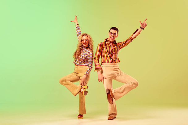 Expressivo, jovem casal talentoso, homem e mulher em trajes vintage elegantes dançando contra gradiente verde fundo amarelo. Conceito de estilo retro, dança disco, moda, arte, hobby, música, anos 70 - Foto, Imagem