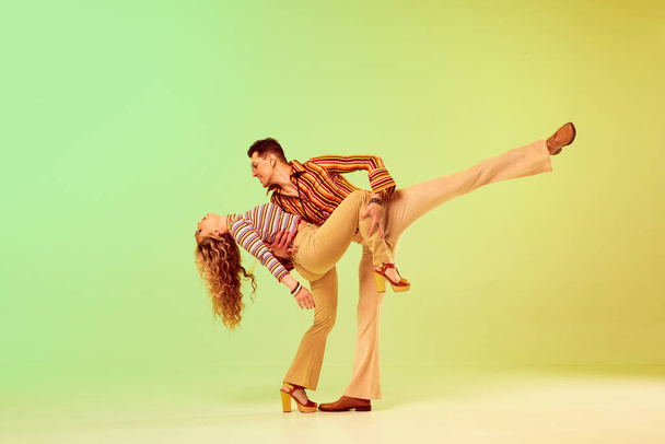 Romantic look. Beautiful young couple, man and woman in vintage stylish clothes dancing disco dance against gradient green yellow background. Concept of retro style, fashion, art, hobby, music, 70s - Foto, Bild