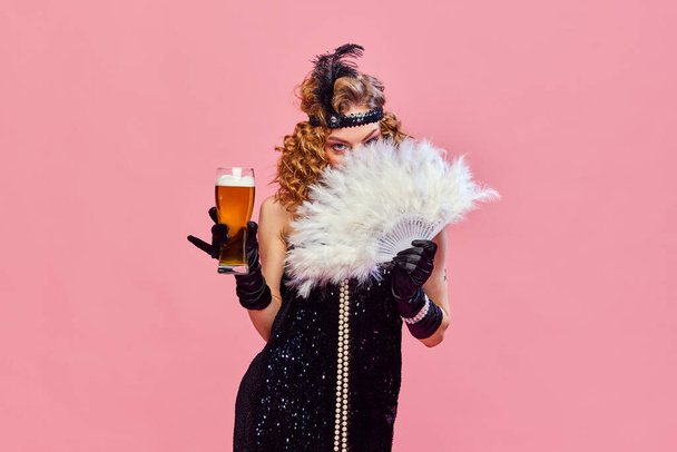 Mysterious woman wearing black vintage dress covers her face with hand fan and holding foamy beer over pink studio background. Concept of drinks, alcohol, Oktoberfest, festivals, rest, vacation, ad - Zdjęcie, obraz