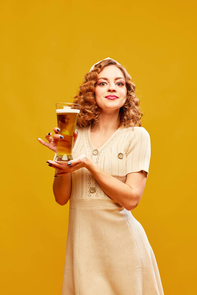 Oktoberfest. Usmívající se sexy dívka, servírka v retro šaty drží pivo sklo a slouží přes žluté pozadí. Koncept nápojů, alkoholu, degustace, festival, dovolená, párty, bavorská kultura - Fotografie, Obrázek