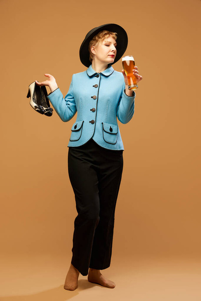 Uma mulher bonita vestindo roupas elegantes com chapéu segurando copo de cerveja sobre fundo bege. Conceito de bebidas, álcool, Oktoberfest, festivais, férias, festa, moda, celebração, anúncio - Foto, Imagem