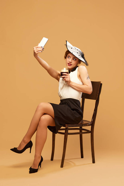 Beer ad. Beautiful woman wearing elegant classical clothes with hat holding glass with alcohol and taking selfie over beige background. Concept of drinks, alcohol, vacation, party, celebrating - Φωτογραφία, εικόνα
