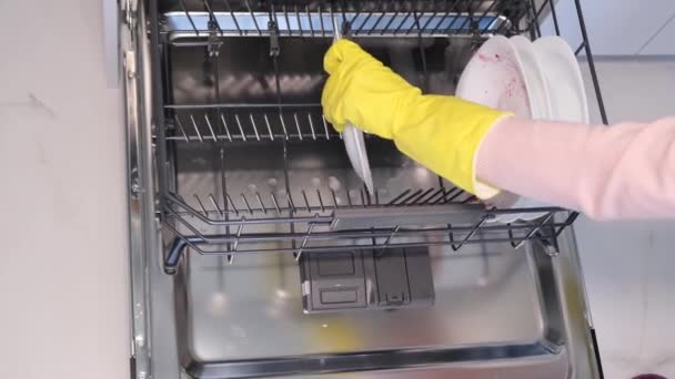Video mano femenina en guantes de goma pone platos blancos en el lavavajillas. Lavado automático de platos sucios en una cocina moderna - Metraje, vídeo