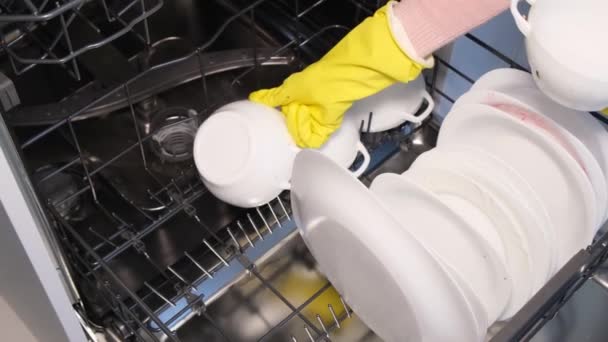 Una mano de mujer con guantes de goma pone platos blancos y tazas en el lavavajillas. Lavado de alta calidad de utensilios de cocina utilizando tecnologías avanzadas - Imágenes, Vídeo