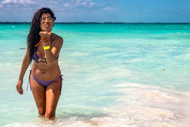 Jong, sexy en mooi meisje in bikini aan de oever van een paradijselijk strand. Toerist geniet van haar reis naar een tropisch strand terwijl ze kusjes met haar handen stuurt. Concept meisje op een strand in een bikini. - Foto, afbeelding